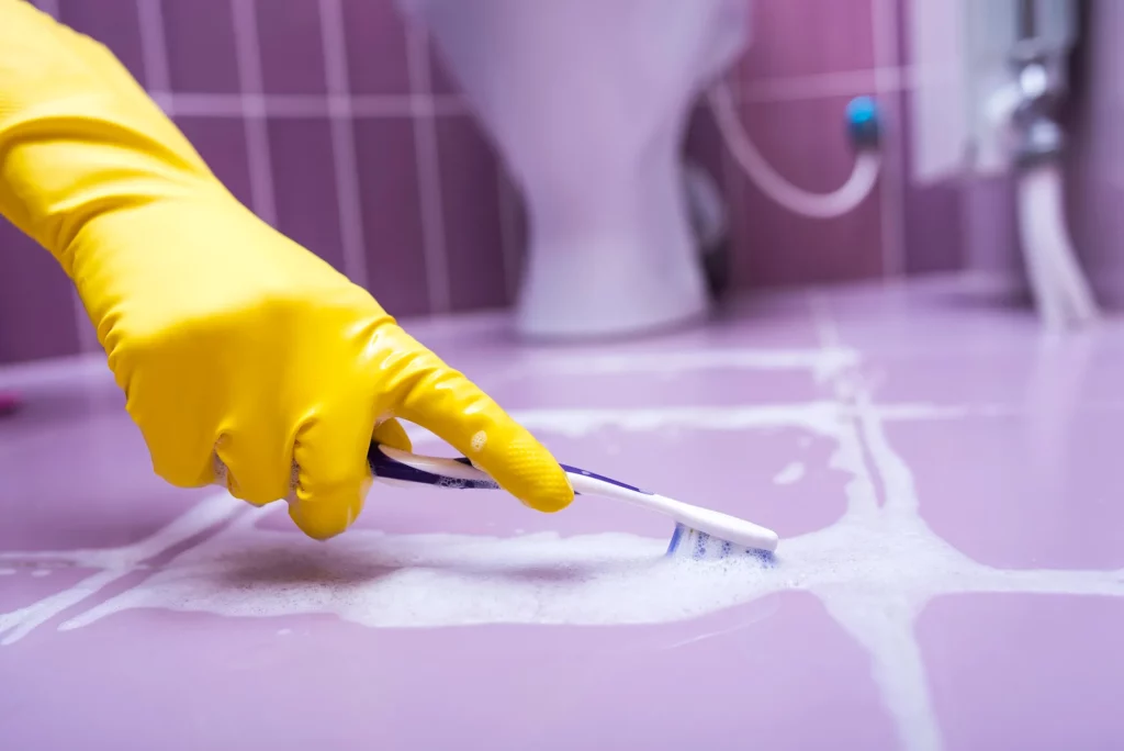Person wearing yellow gloves cleaning bathroom tile grout with a toothbrush
