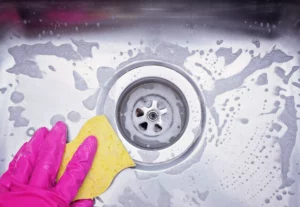 person wearing pink rubber gloves scrubbing a stainless steel kitchen sink drain with a yellow sponge. Cleaning drains inside